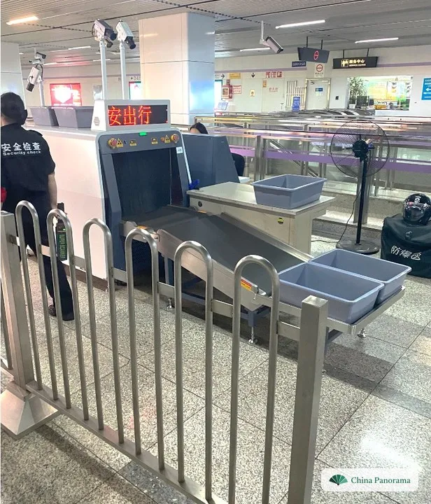 Security check in a metro station: X-ray machine with conveyor belt and grey plastic trays, with a barrier in front