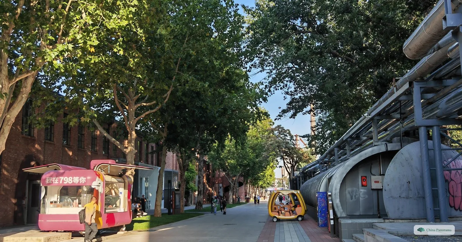 A tree-lined street in the 798 Art District, with old industrial pipes running overhead on the right and a pink snack cart on the left