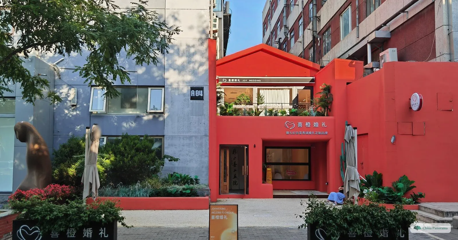 A bright red low building with a terrace among grey blocks in the 798 Art District, with a sculpture and flower beds in front