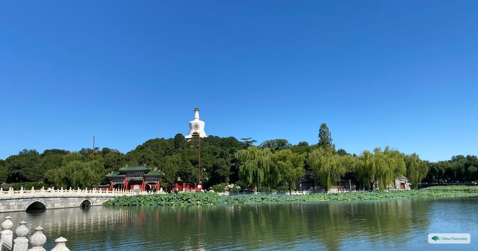 The White Dagoba on its wooded island in Beihai Park, seen across the lake with a marble bridge on the left and lotus along the shore