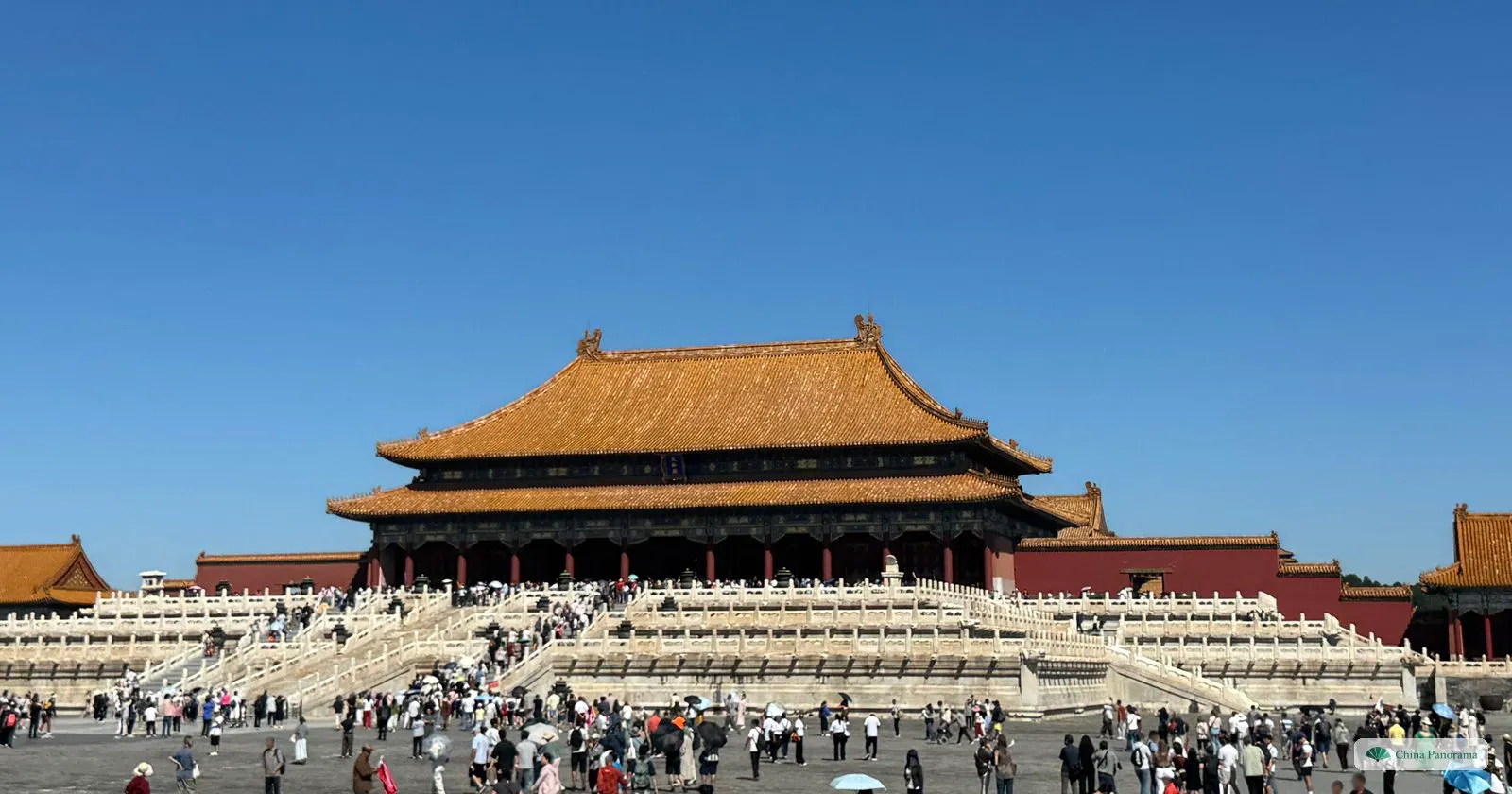 The Hall of Supreme Harmony in the Forbidden City on its three-tiered white marble terrace, under a deep blue sky, with visitors crossing the courtyard in front