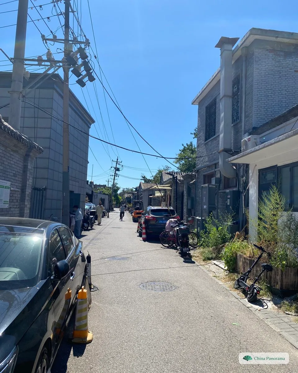 A Beijing hutong at midday: a narrow lane between grey-brick houses with parked cars, overhead cables and a bicycle against a wall