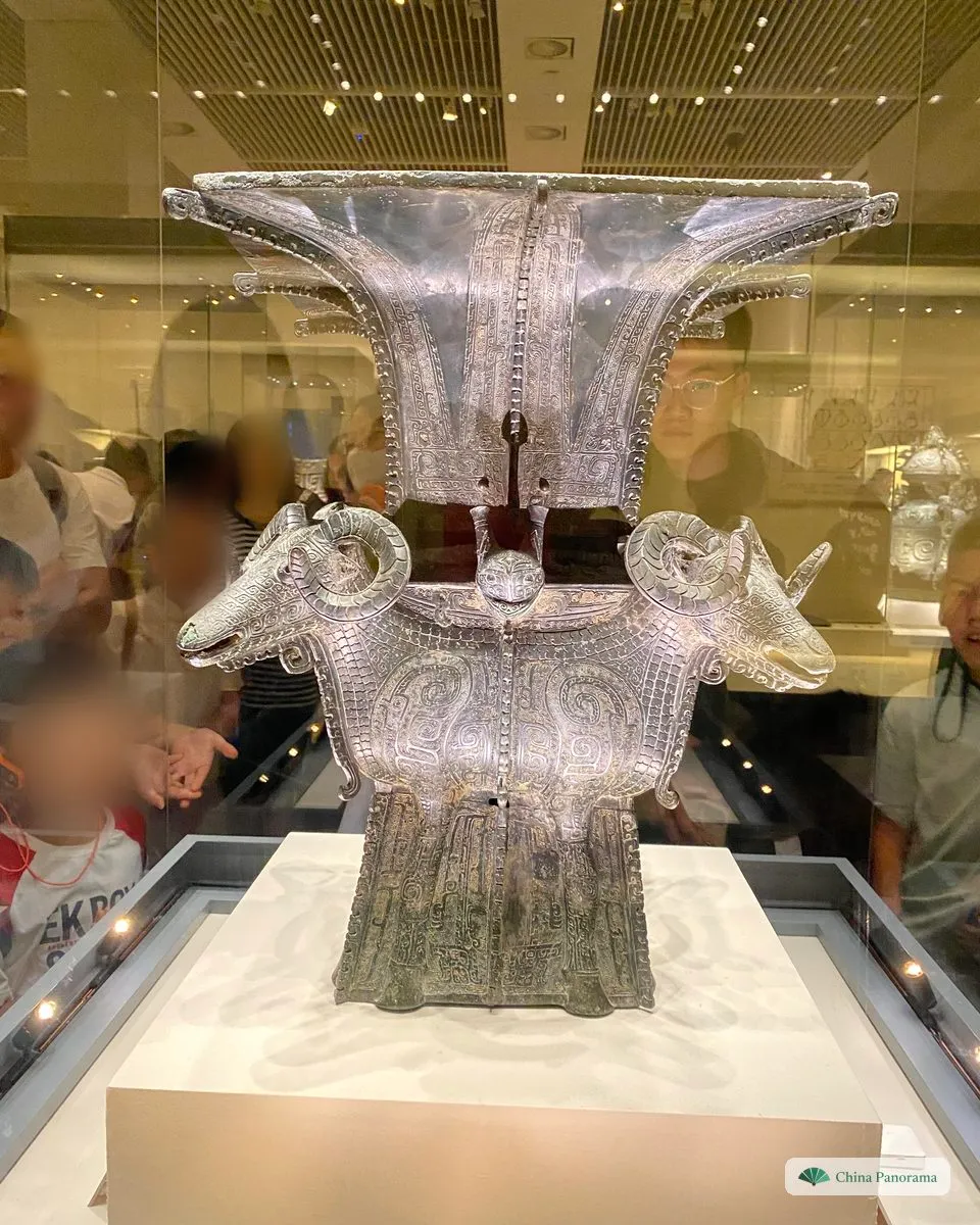 A Shang-dynasty bronze wine vessel in a museum showcase: a square zun with a flaring rim and a ram’s head with curled horns at each corner