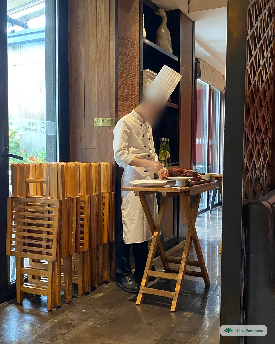 A cook in a tall white hat carving a roast Peking duck at a wooden trolley beside the tables of a restaurant