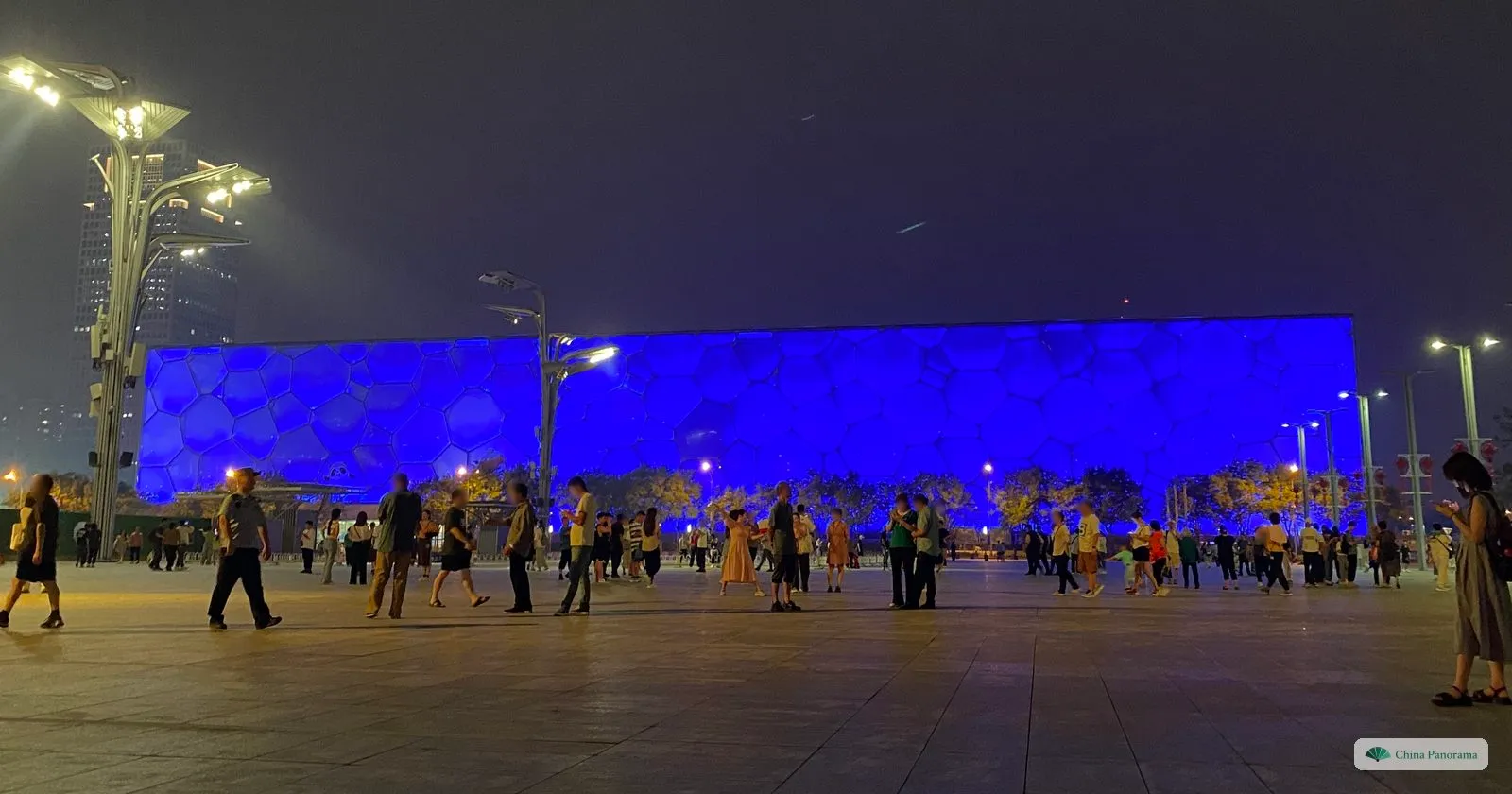 The National Aquatics Center, the Water Cube, glowing blue at night, with people strolling across the plaza in front