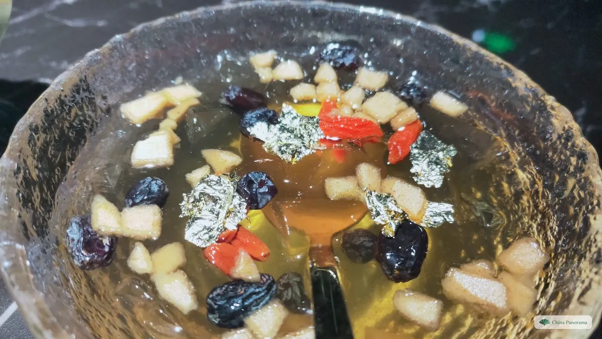 A glass bowl of clear, golden jelly in syrup with diced apple, raisins, goji berries and flakes of gold leaf, a spoon in it