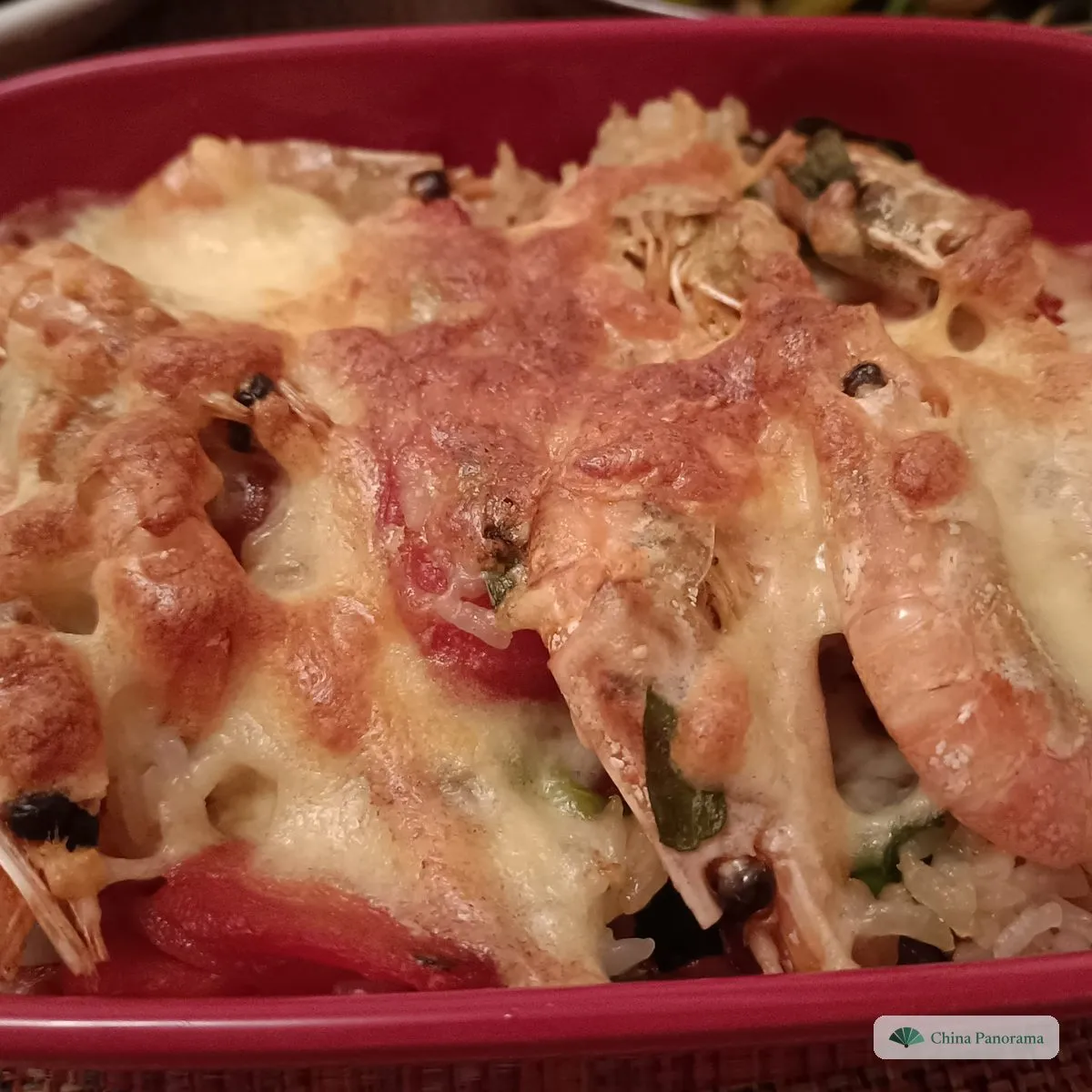 A red oval baking dish of rice under prawns, tomato and browned melted cheese