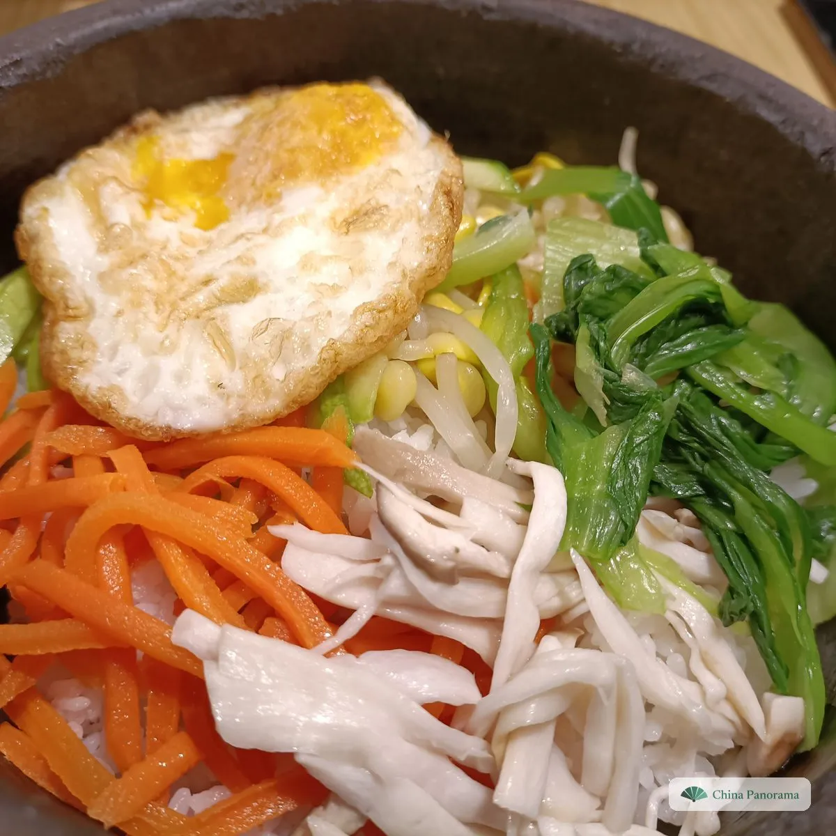 A stone bowl with a fried egg, carrot strips, bean sprouts, greens and white mushroom strips laid in sections over rice