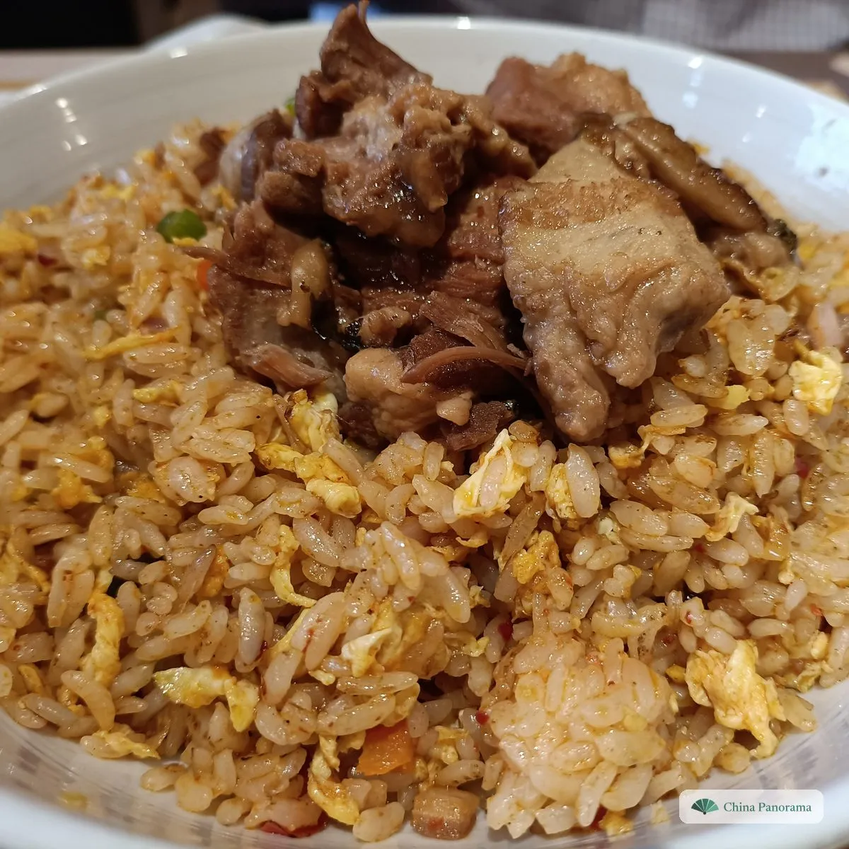 A white bowl of golden fried rice with scraps of egg and flecks of chilli, heaped with chunks of braised pork ribs and pork belly