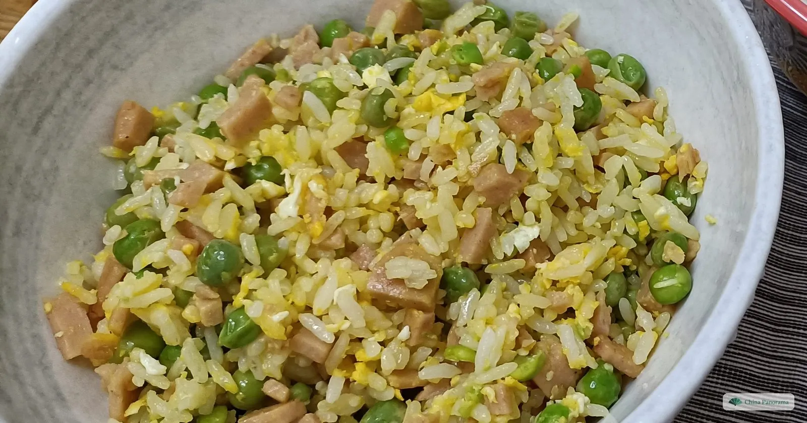 A grey-white bowl of pale gold fried rice, close up: separate grains with soft scraps of egg, pink cubes of ham and bright green peas