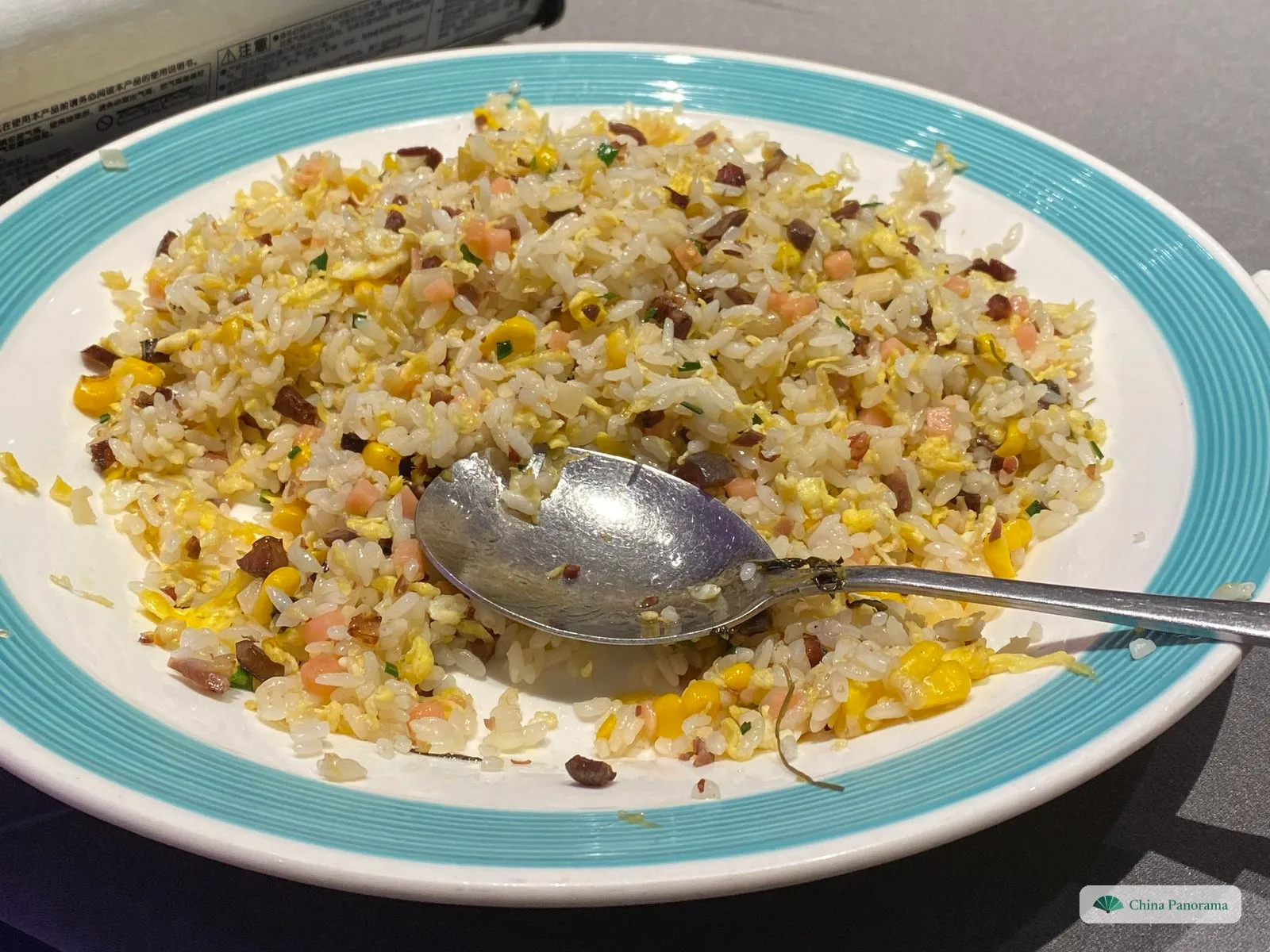 A white plate with a turquoise rim, half covered in pale fried rice with yellow egg, pink and dark brown cubes, sweet corn and flecks of spring onion; a metal serving spoon lies in it
