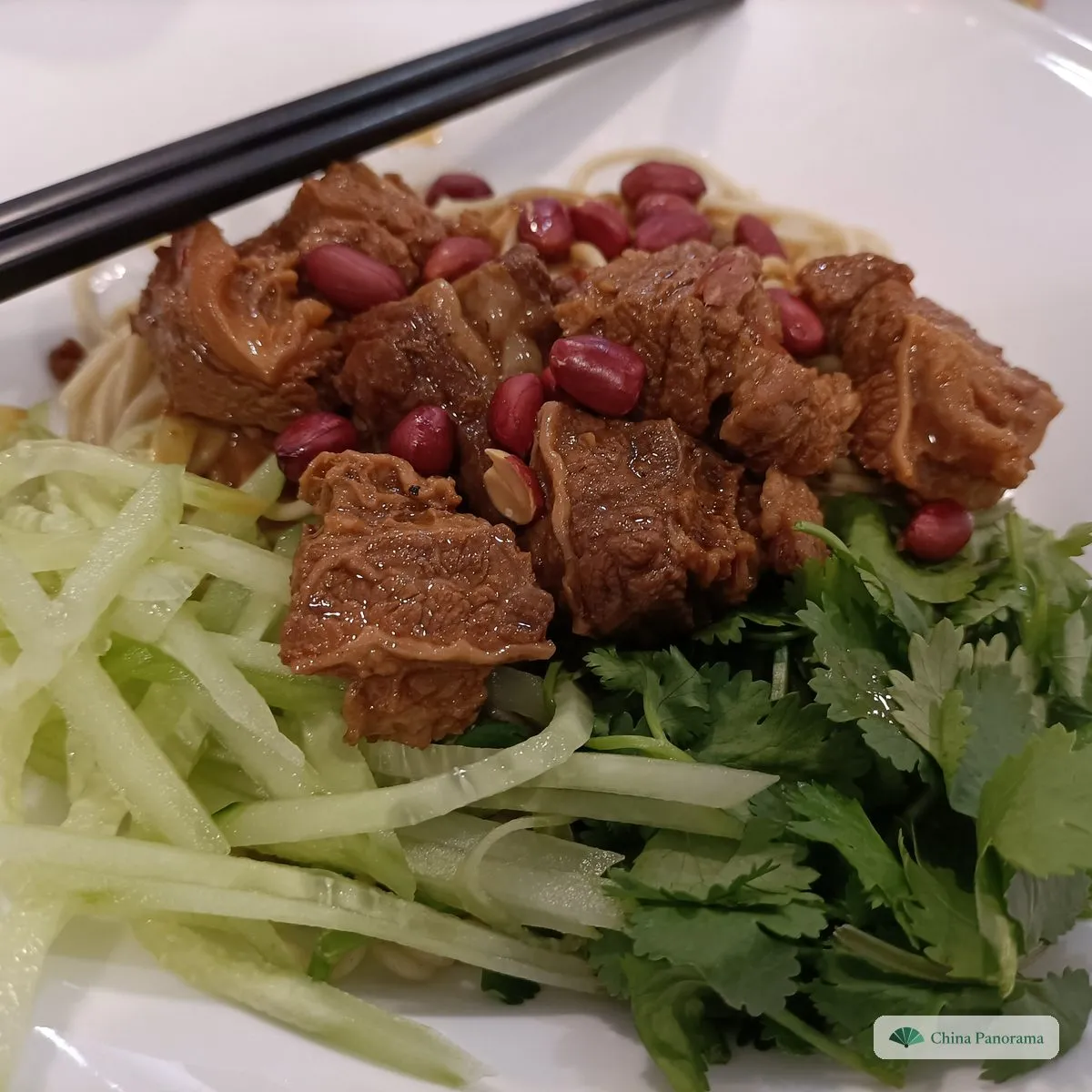 Noodles covered with chunks of braised beef, red-skinned peanuts, shredded cucumber and coriander, chopsticks on top