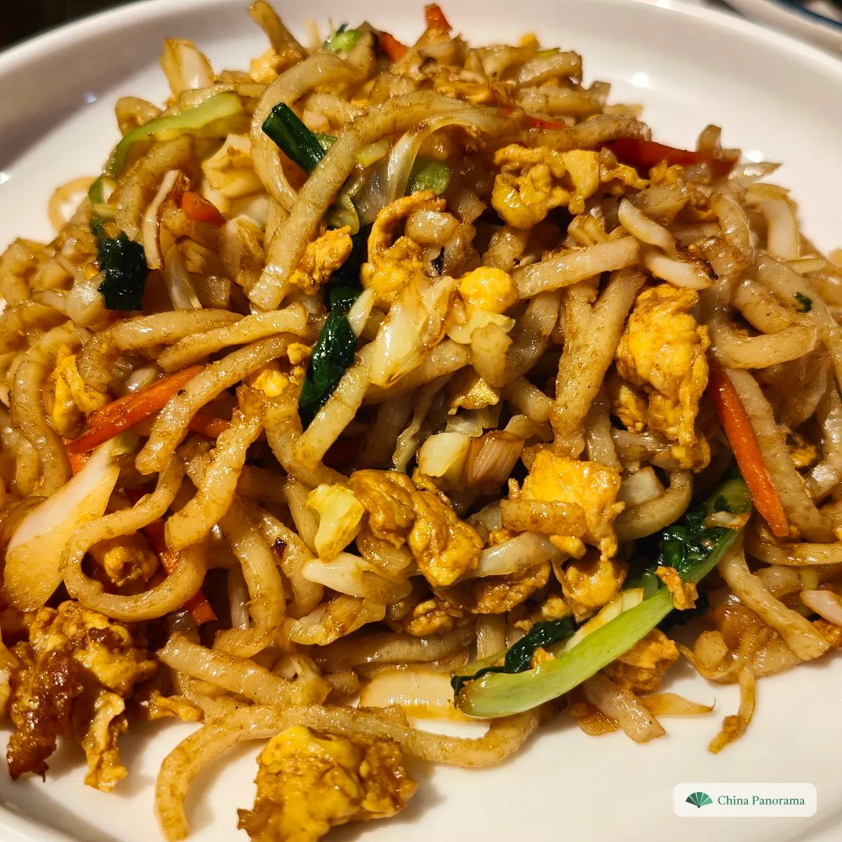 A white plate of pale, uneven fried noodles with scrambled egg, cabbage, carrot strips and greens