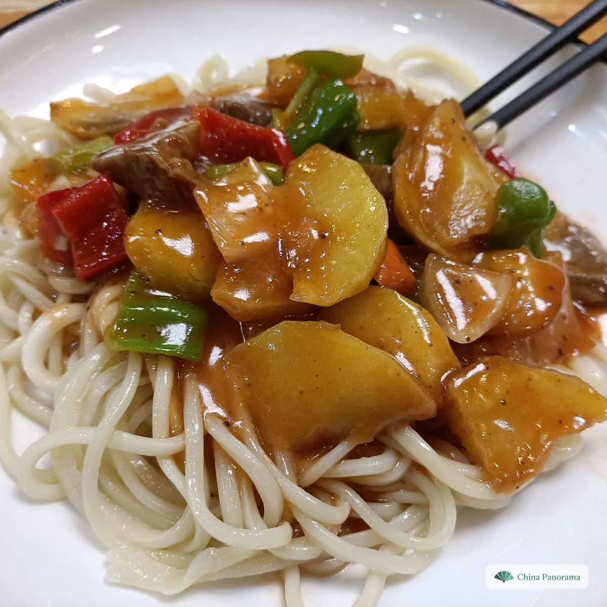 Boiled white hand-pulled noodles on a plate under potato chunks, red and green peppers and meat in a glossy brown sauce, chopsticks at the side
