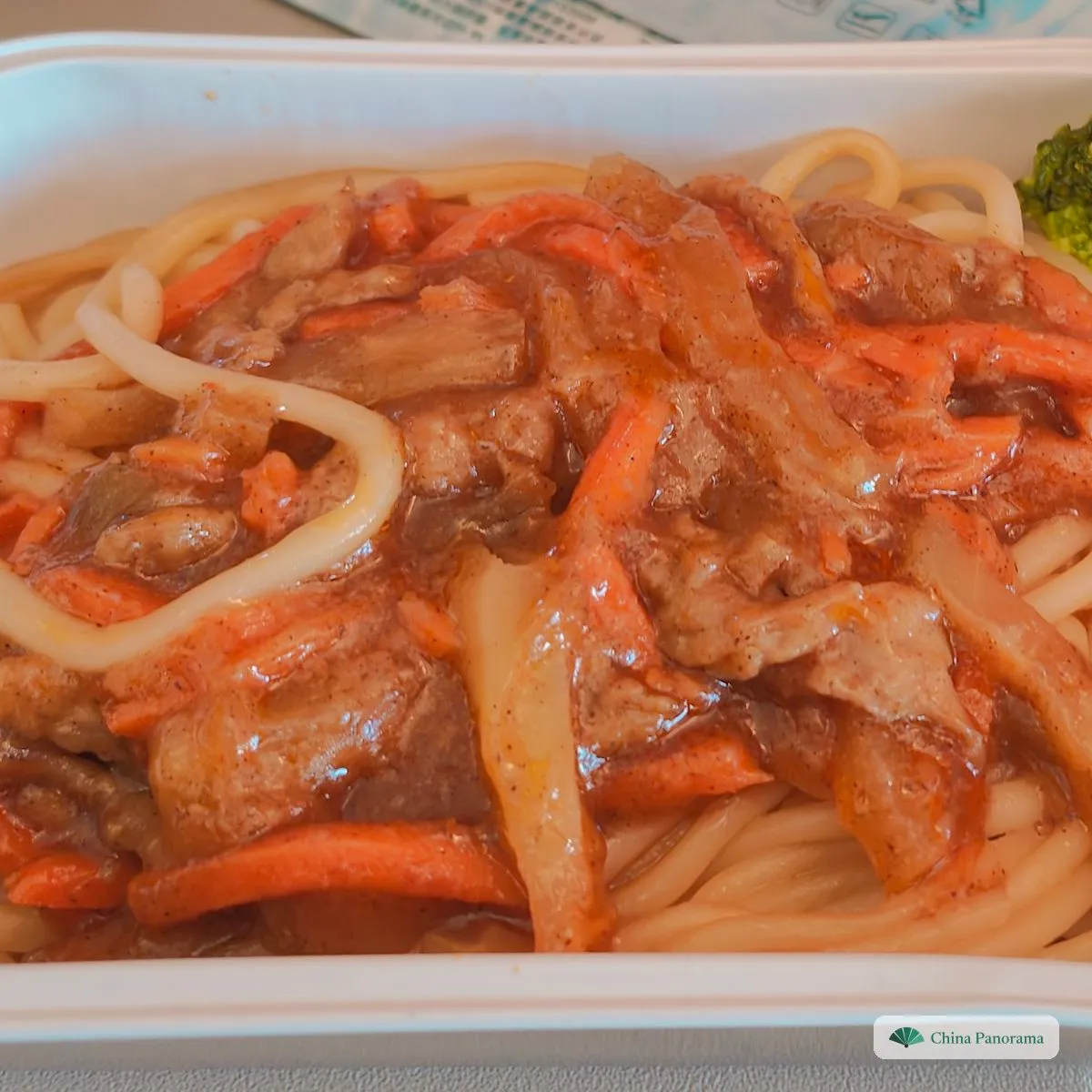 Thick spaghetti in a plastic box under strips of beef, onion and carrot in brown sauce, a floret of broccoli in the corner
