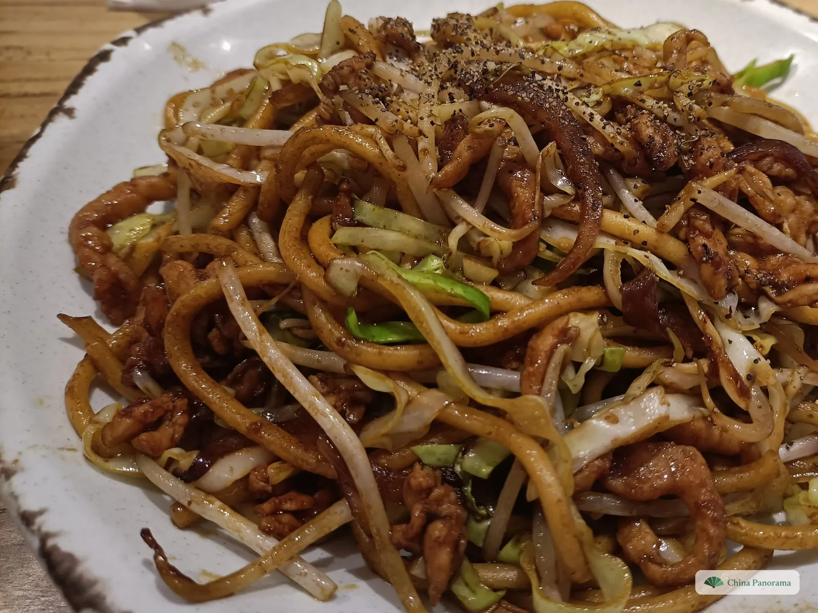 A close-up of a white plate of thick, soy-brown noodles with shredded pork, bean sprouts, pale green cabbage and ground black pepper