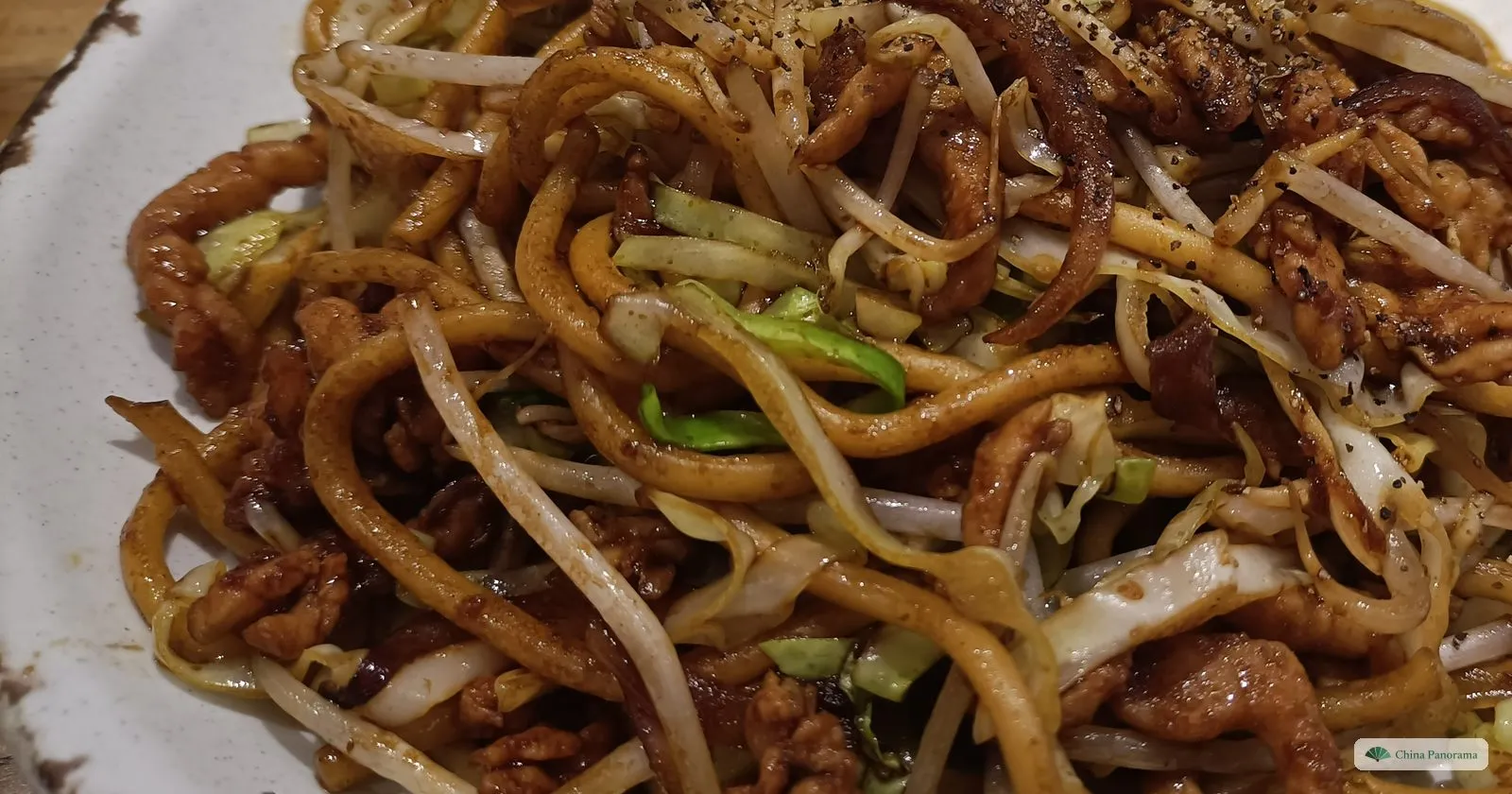 A white plate heaped with thick, soy-brown noodles, shredded pork, bean sprouts and pale green cabbage, dusted with black pepper