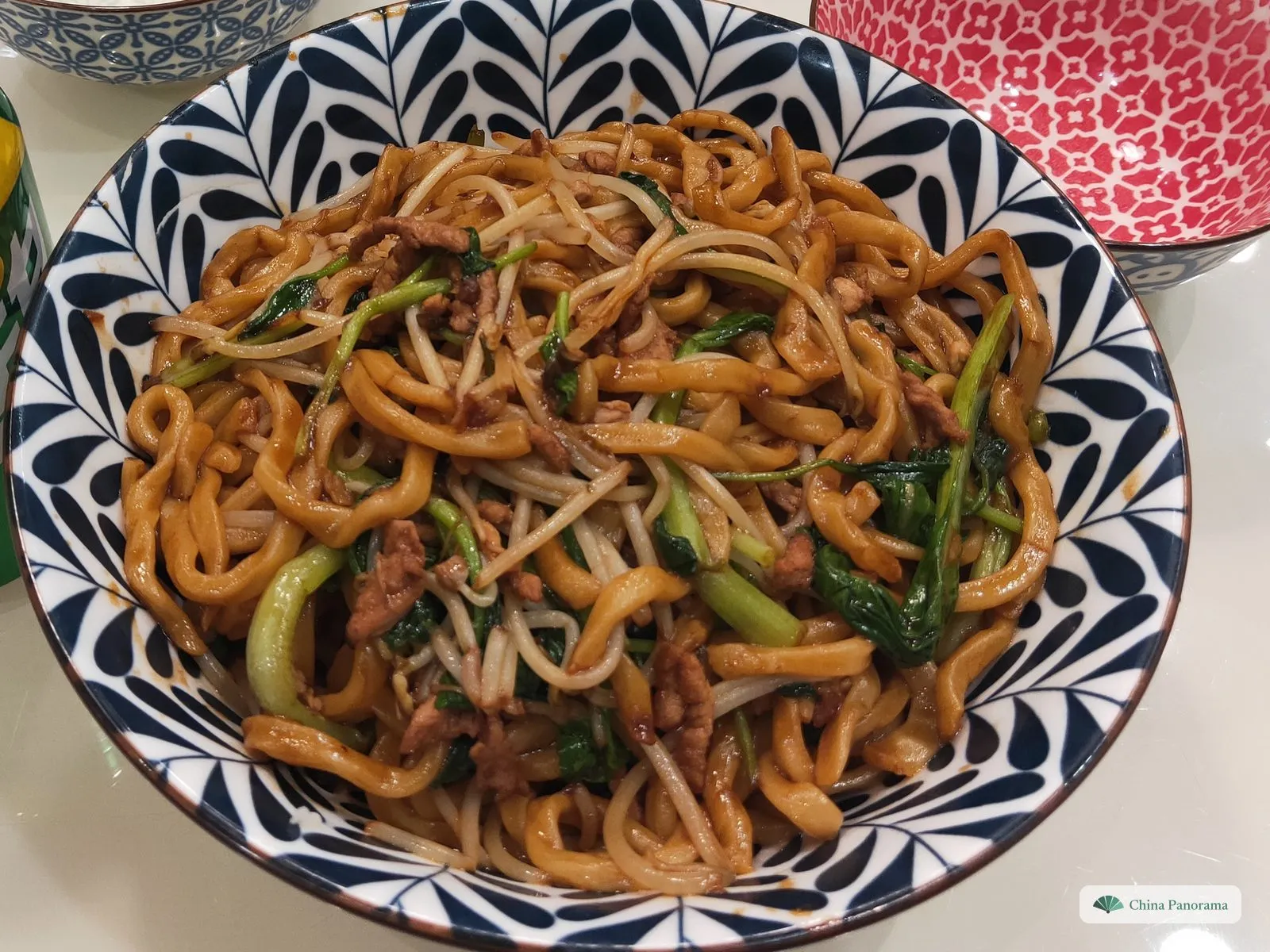 A blue-and-white bowl seen from above, full of thick, glossy brown noodles with shredded pork, bean sprouts and dark green leafy greens