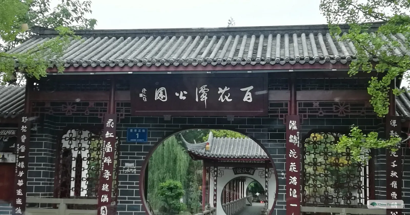 The gate of Baihuatan Park: a dark brick wall with a round moon door, a black plaque 百花潭公園 and calligraphy on red boards, a covered walkway seen through the door