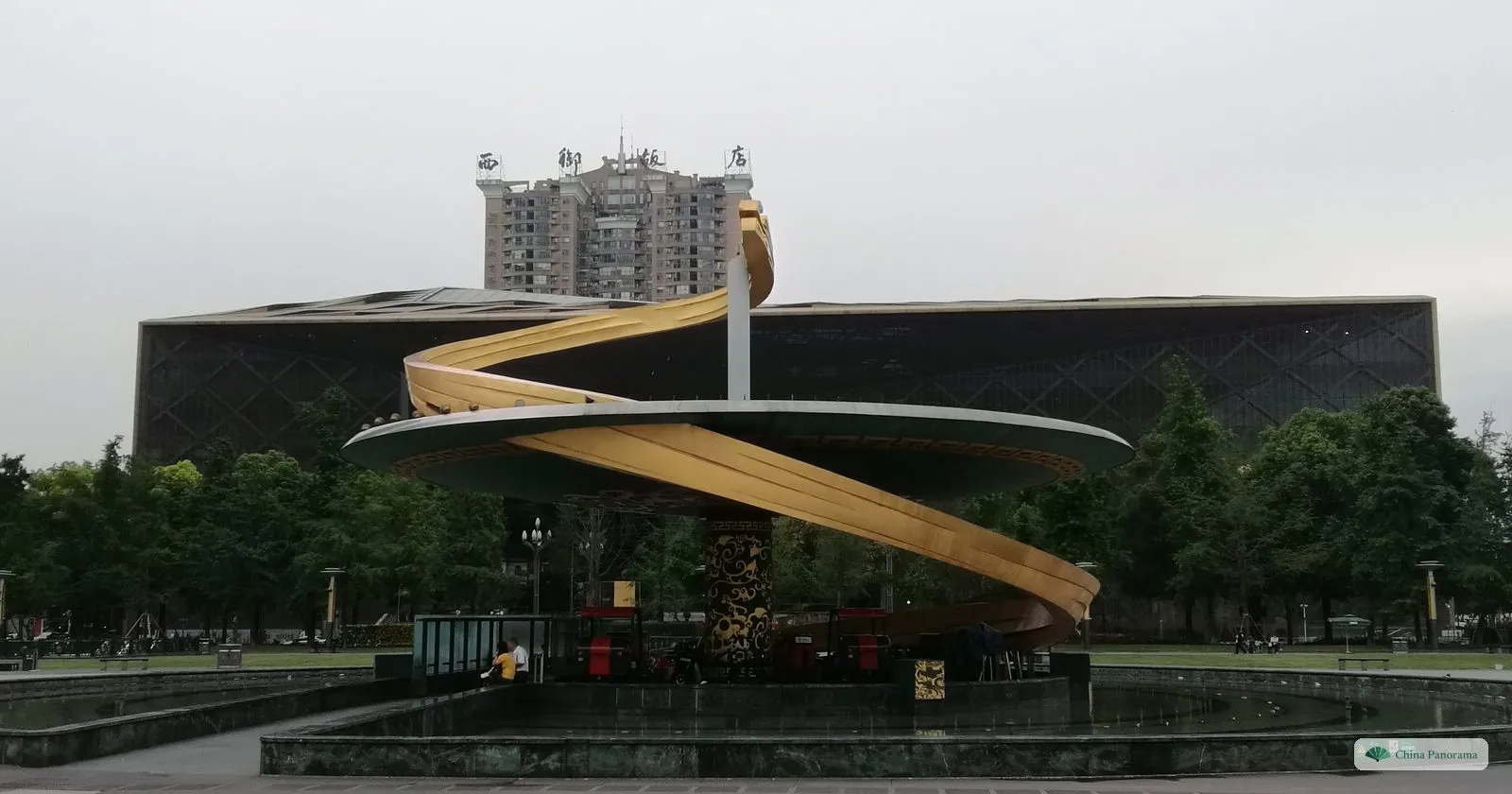 The Chengdu Museum: a long dark building wrapped in a diamond lattice, behind a round fountain basin with a golden spiral sculpture on the square