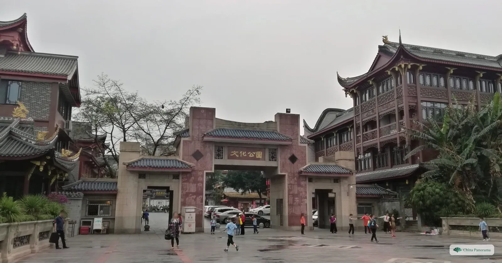 The pink stone gate of the Culture Park with the plaque 文化公园, tall red-and-brown traditional buildings either side and people exercising on the wet square