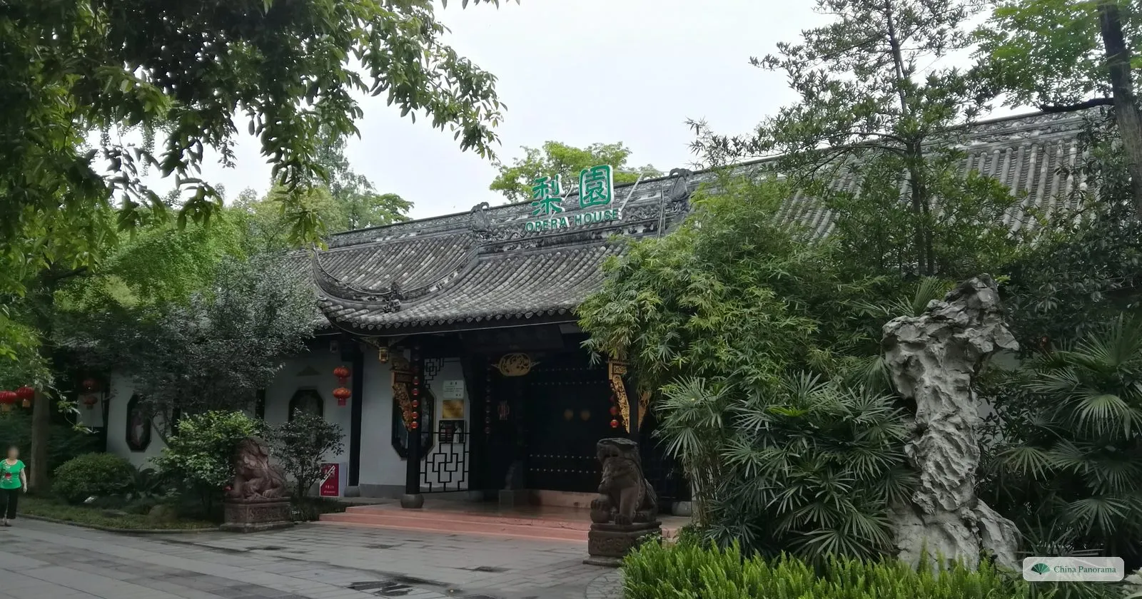 A traditional hall with a grey tiled roof and the green sign 梨園 Opera House, a stone lion, a scholar’s rock and bamboo in front
