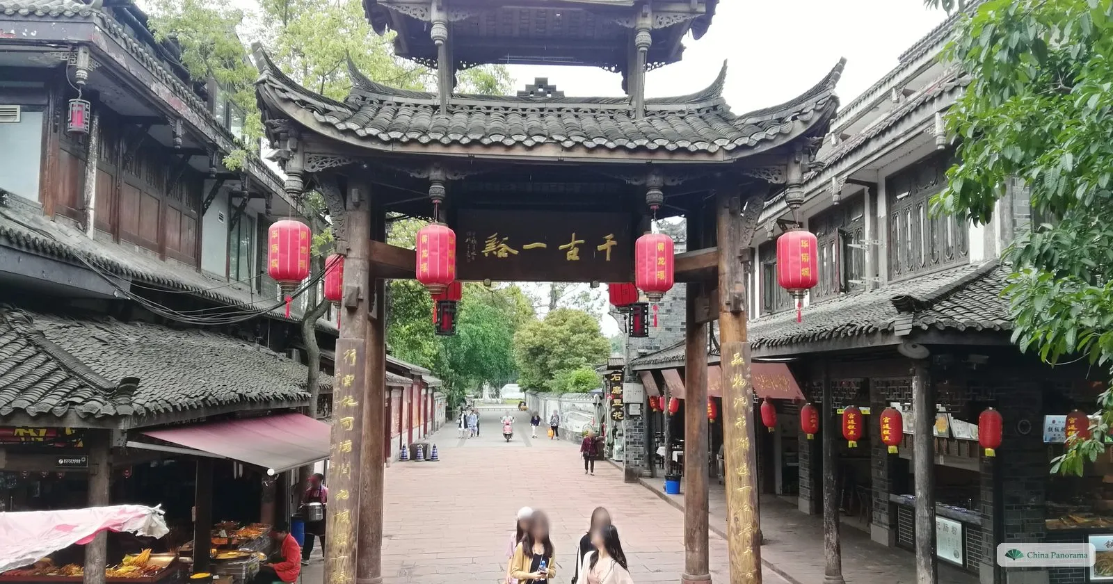 A wooden archway with a tiled roof and red lanterns over a paved street, the plaque reading 千古一绝, wooden shopfronts either side