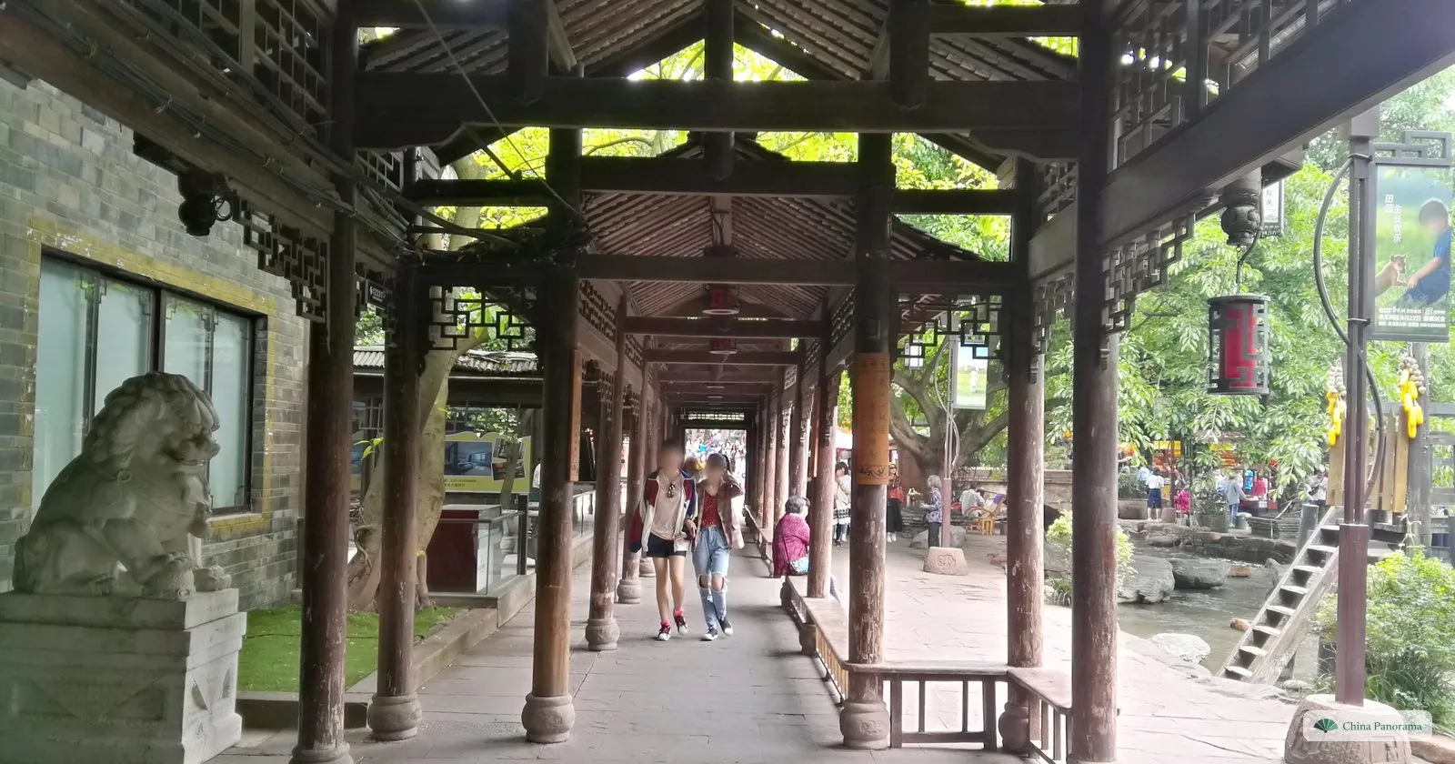 A long covered wooden walkway with dark pillars and carved brackets, a stone lion at its start and visitors walking along it, trees and a stream to the right