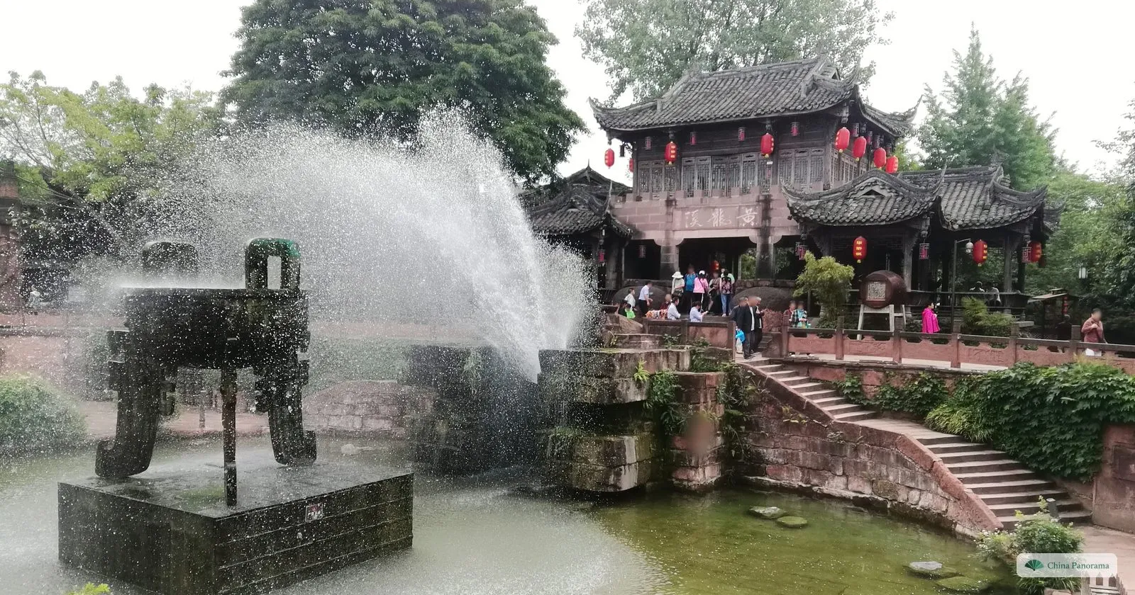 A large bronze tripod vessel standing in a pool, a fountain spraying beside it and a wooden pavilion with red lanterns on the far bank