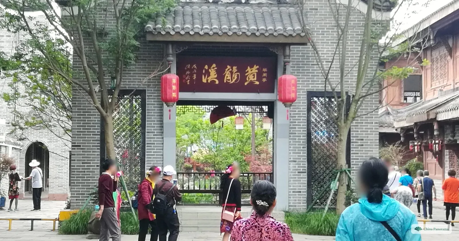 A grey brick gate with a red plaque 黃龍溪 and red lanterns, a garden visible through it and tourists in front