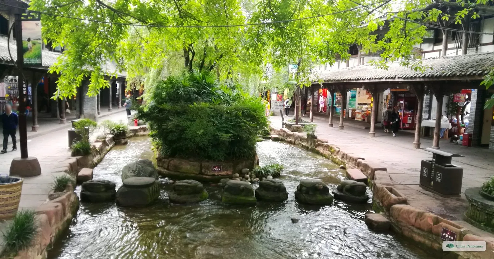 A clear stream running between stone banks through a little town, with a row of round stepping stones, a green island and covered arcades of shops on both sides
