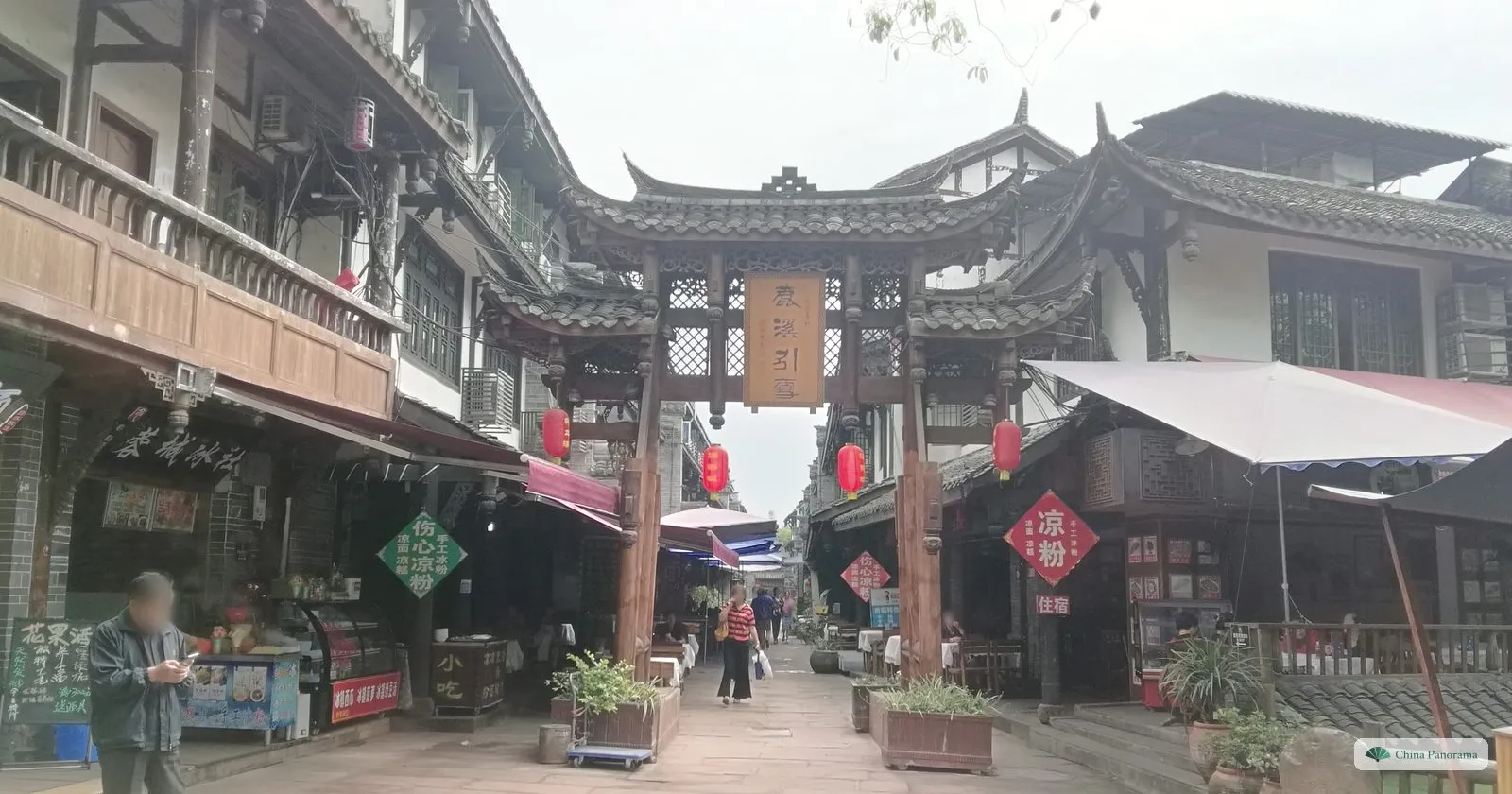 An old street of two-storey wooden houses with a carved wooden archway, red lanterns, small shops and a few people