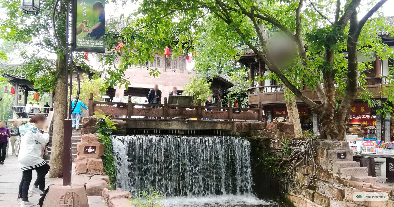 A small waterfall pouring over a stone weir under a balustrade, with green trees, roots over the rocks and visitors on the paths