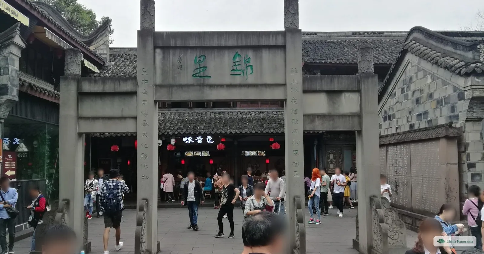 A plain grey stone archway with green characters carved on the lintel, a snack restaurant behind and people walking through