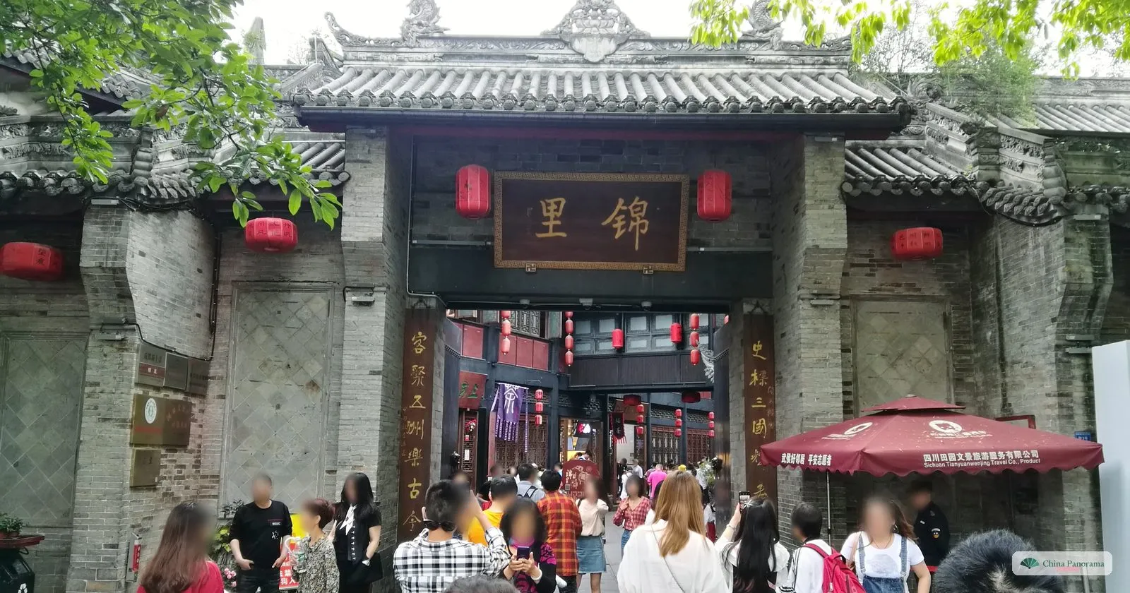 The gate of Jinli: grey brick walls with a wooden plaque 锦里, red lanterns, a red umbrella and a crowd going in