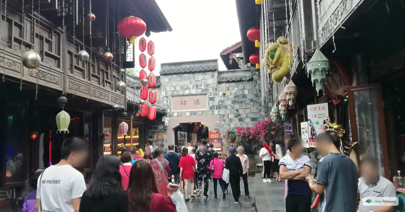A packed lane between dark carved wooden shopfronts, strings of red lanterns and glass baubles overhead, a brick gate tower at the end and a poster for a face-changing opera show