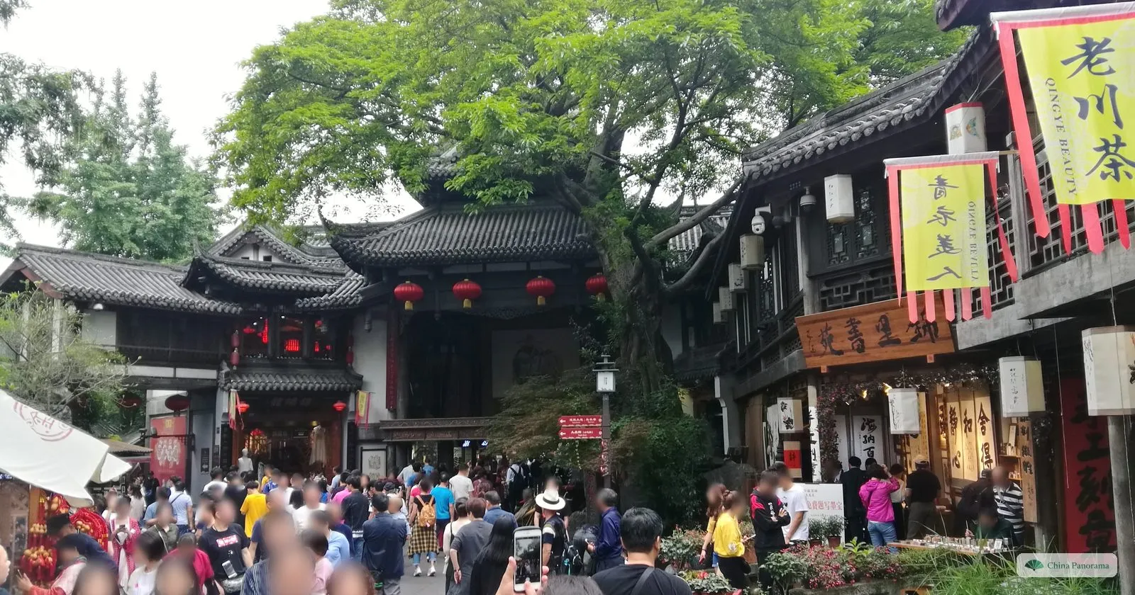 A small square under a huge old tree, surrounded by two-storey wooden buildings with upturned eaves, yellow banners and red lanterns, filled with people