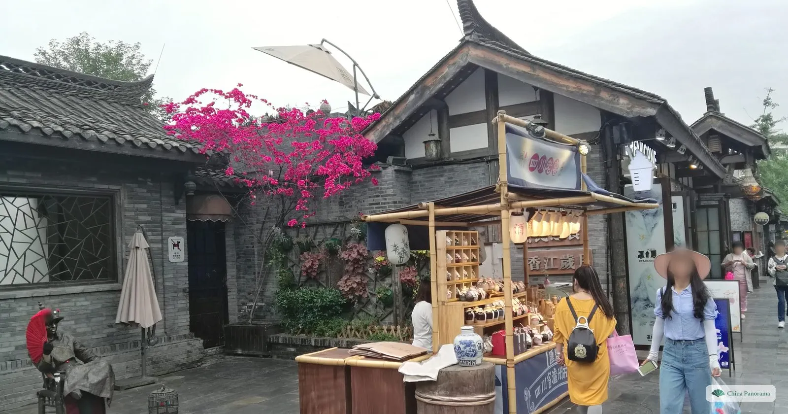 Pink bougainvillea over a grey brick wall and a round window, a bronze figure of a man with a fan by a doorway, and a bamboo stall selling small jars