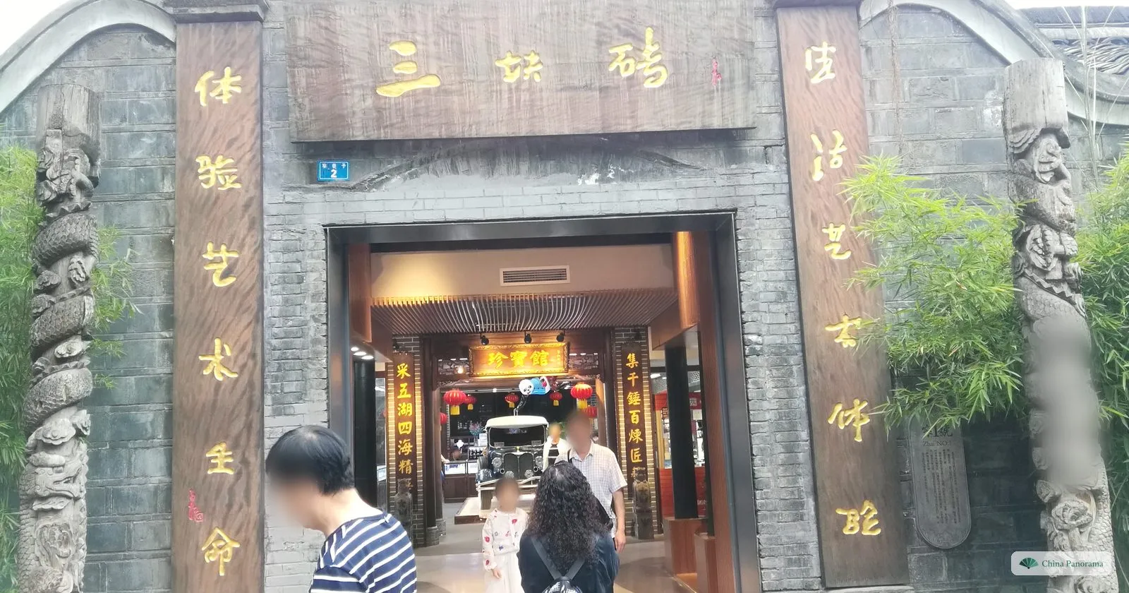 A courtyard gate set in a grey brick wall, with carved stone pillars, a wooden board with three gold characters and vertical gold inscriptions either side, a shop inside