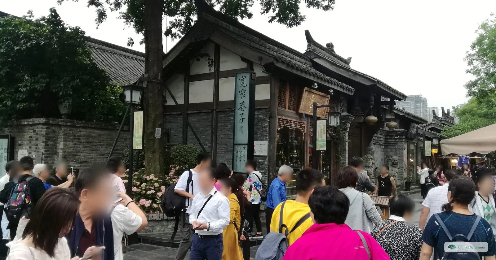 A tall green signpost reading 宽窄巷子 in front of a house with a dark timber frame and whitewashed panels over a grey brick base, a crowd of visitors in front