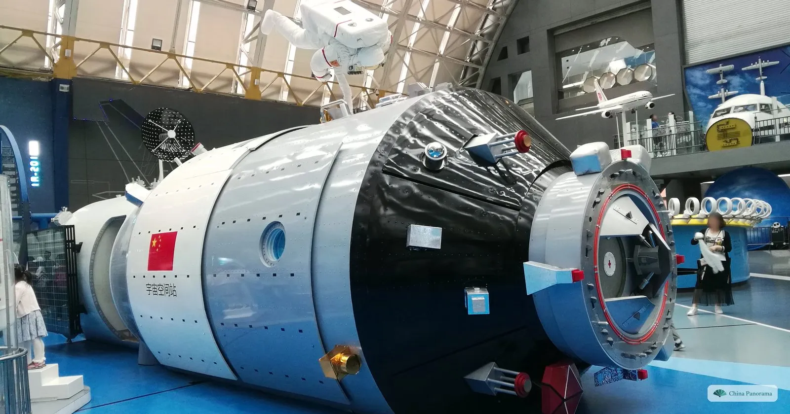 A large model of the Tiangong space-station module with a Chinese flag, inside a hall with aircraft models and an astronaut figure overhead