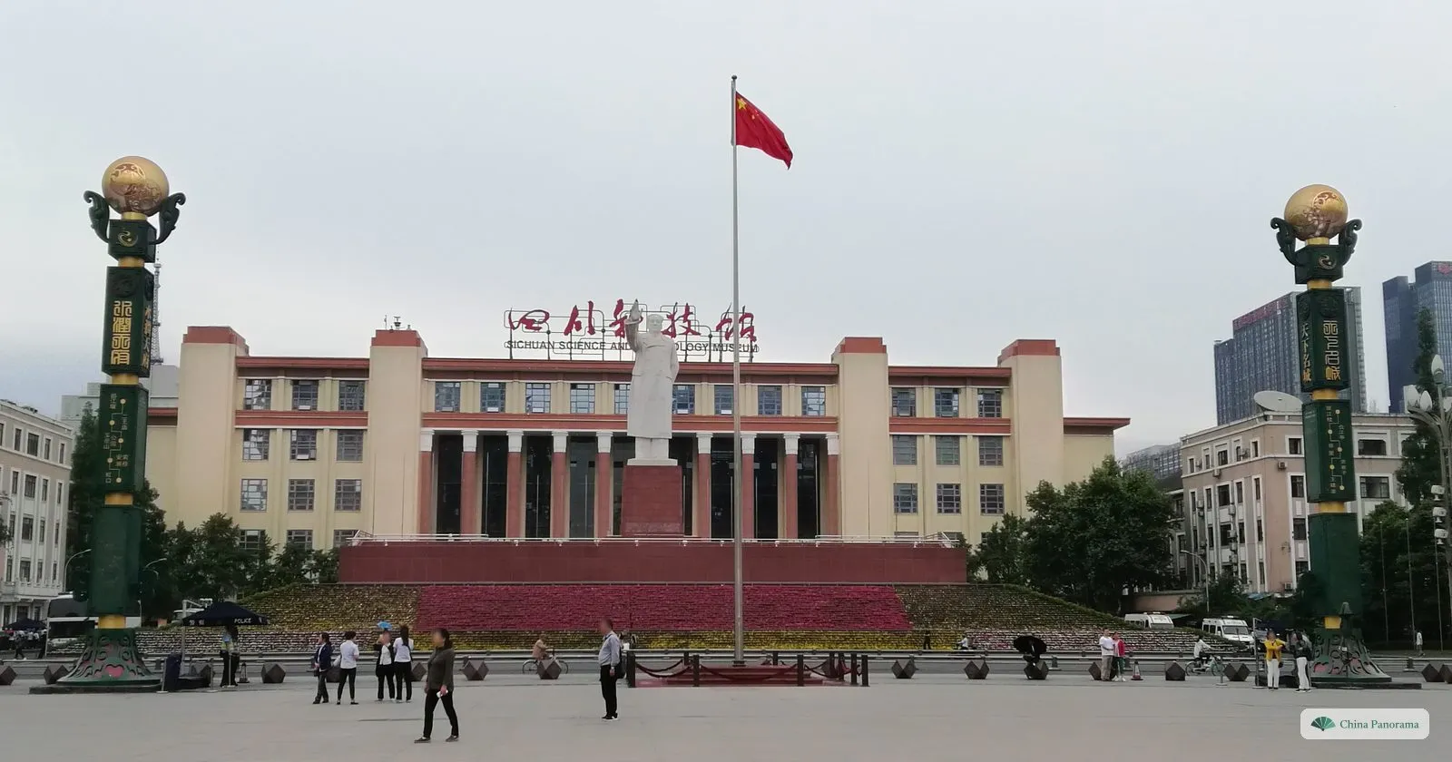 The Sichuan Science and Technology Museum: a long cream building with red columns and the red sign 四川科技馆, the Mao statue and a flag in front, green columns topped by gold globes either side