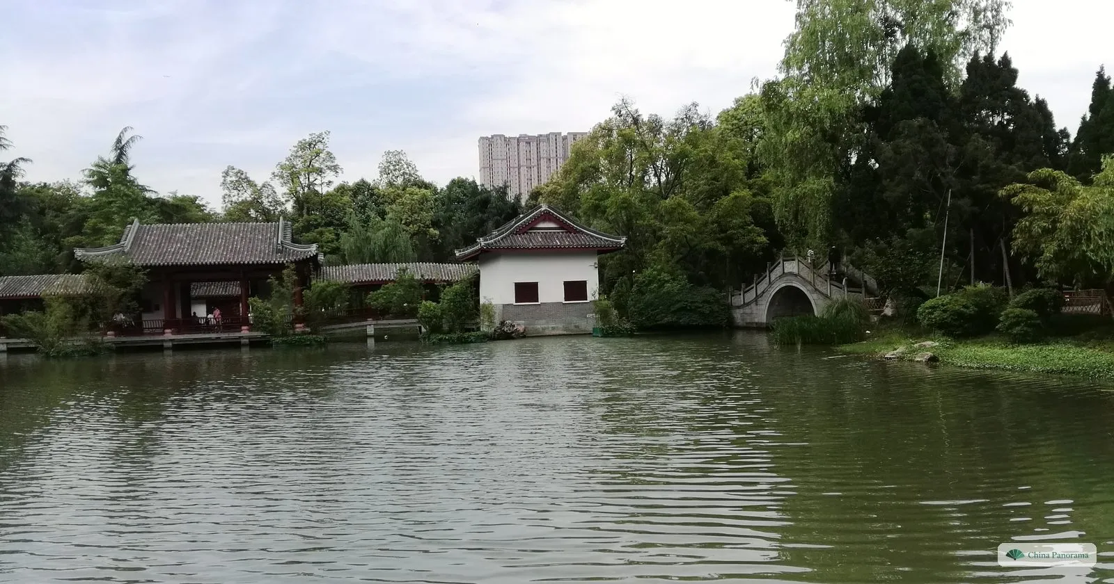 A quiet lake in the garden of the Wuhou Shrine with a white-walled pavilion, a red-pillared hall, a small arched stone bridge and dense trees