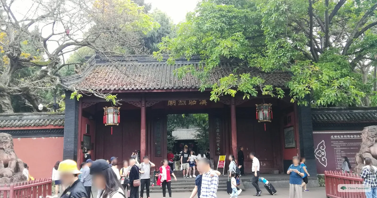 The red main gate of the Wuhou Shrine under big trees, with a gold plaque 漢昭烈廟 over the doorway, stone lions either side and visitors on the steps