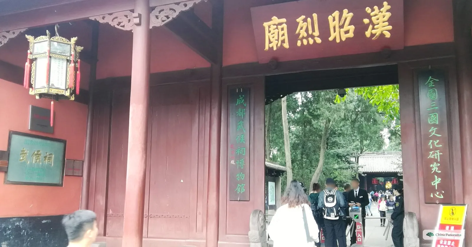 Close view of the shrine gate: the gold characters 漢昭烈廟 on a red board above the door, vertical green inscriptions on the posts, a hanging lantern and a small green plaque reading 武侯祠 on the wall to the left