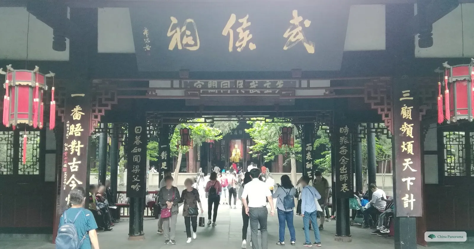 A wooden passage hall with the plaque 武侯祠 in flowing calligraphy and couplets on the pillars, looking through to a gilded statue in the hall beyond, visitors walking through