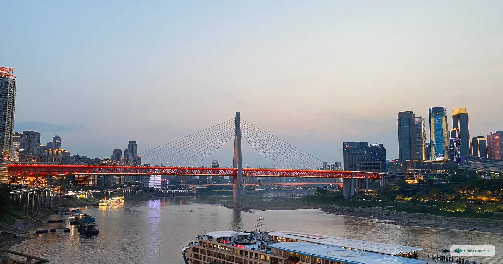 The Jialing River at dusk from Chaotianmen: the red truss and white tower of the Qiansimen Bridge, office towers on the right bank and a cruise ship in the foreground