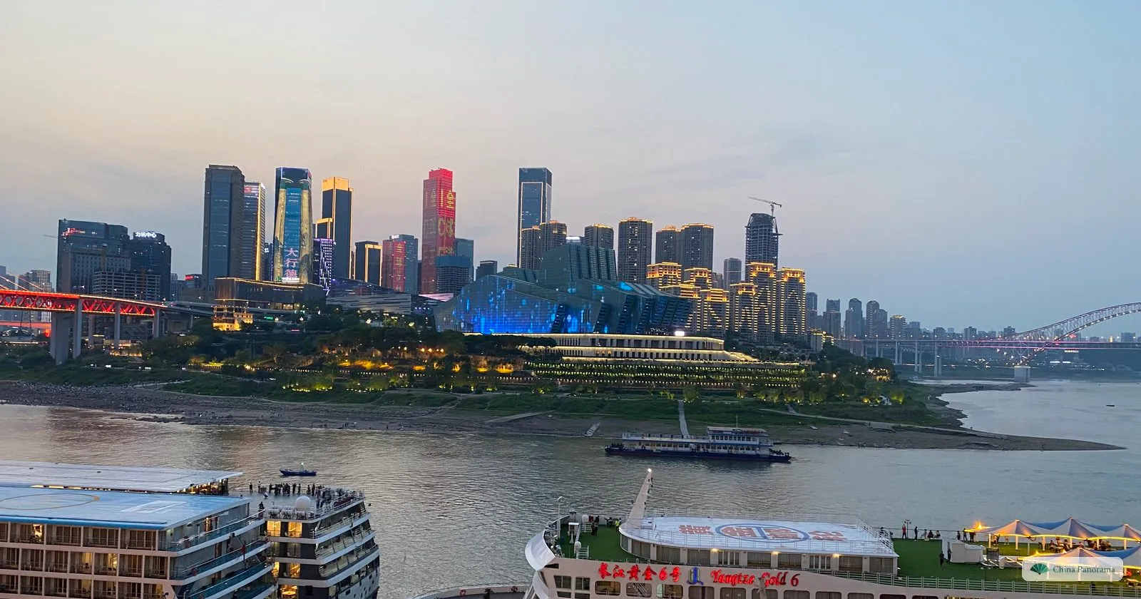 Dusk at the Chaotianmen piers: two Yangtze cruise ships moored below, the lit towers of Jiangbeizui across the river, the blue-lit Grand Theatre on the point and an arch bridge in the distance