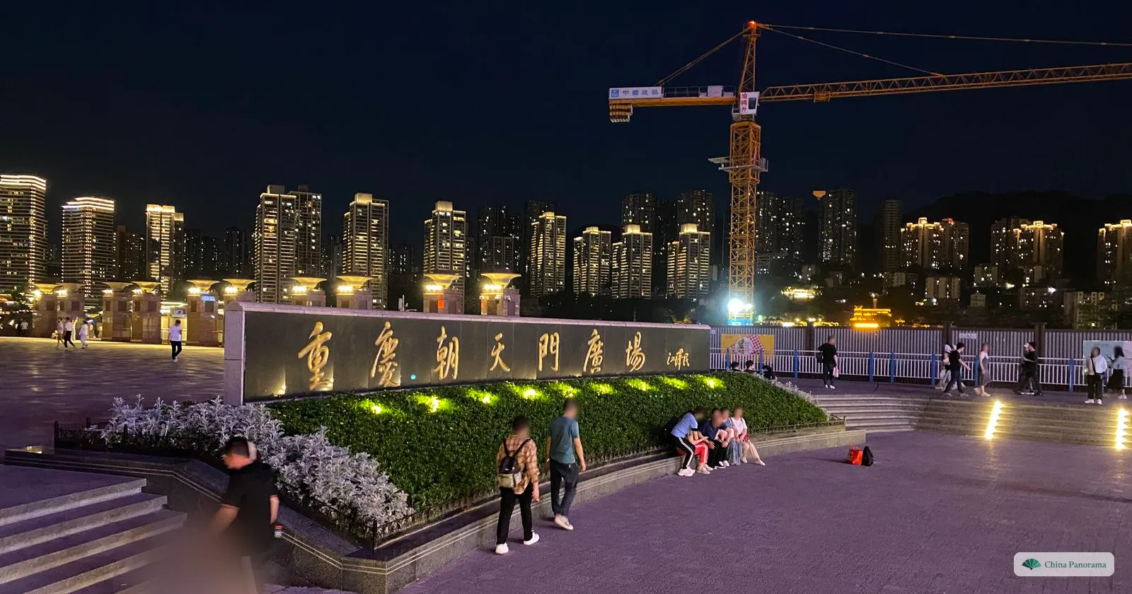 Chaotianmen Square at night: a long stone carved with 重庆朝天門廣場, flower beds and steps, a few people walking, lit apartment towers and a crane behind