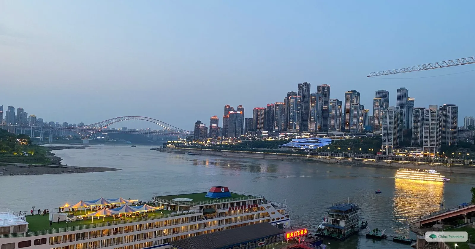 The Yangtze at dusk looking downstream from Chaotianmen, an arch bridge on the horizon, high-rise flats on the far bank and a lit tour boat on the water